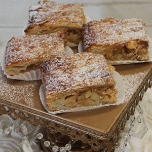 Apple strudel slices with golden, flaky pastry and a dusting of powdered sugar are displayed on decorative parchment atop an ornate gold cake stand.