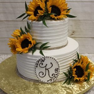 The TC-006 is a two-tiered white cake adorned with large sunflowers, green leaves, and a brown “K” monogram on the lower tier, all displayed on a gold textured cake board.