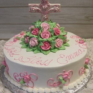 The RC-085A is a round white cake with pink and green icing roses, an ornate pink cross topper, “God Bless Emma” in pink piping, and floral accents on the sides.