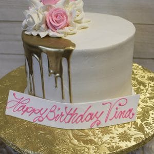 The BD-057C Gold Drip Partial is a white cake with gold drip icing, pink and white icing roses on top, on a gold floral-patterned board. A "Happy Birthday Tina" sign in pink script sits in front.