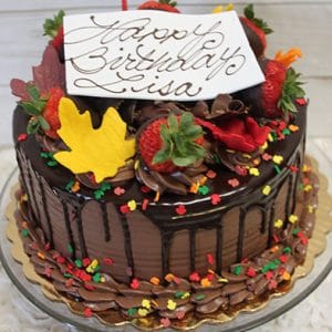 The THX-013 is a chocolate birthday cake topped with strawberries, autumn leaf-shaped decorations, sprinkles, and a white plaque reading "Happy Birthday Lisa" in cursive. The cake is displayed on a glass stand.