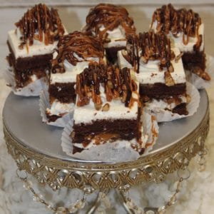 Six Turtle Squares topped with white frosting and caramel drizzle are displayed on an ornate silver cake stand with decorative chains, set against a textured white background.