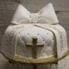 The RC-061 cake is a round white cake featuring quilted detailing with small gold accents, a large fondant bow, a gold cross, and gold writing that says "God Bless" and "Christiano.