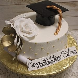 GC-045: White round cake with a large white rose, gold flower, gold pearl accents, and a black graduation cap with gold tassel. “Congratulations Dilma” sign on top.