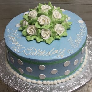 A round blue BD-160 cake with white polka dots, green trim, and "Happy Birthday Maureen" in white icing. Topped with white icing roses and leaves, it sits on a silver foil-covered board.
