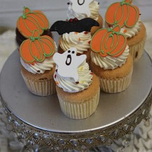 Six CC-052 Mini Halloween cupcakes Gold with chocolate mousse filling are topped with festive decorations like fondant pumpkins, ghosts, and a bat, and beautifully arranged on an ornate silver cake stand.