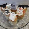 Four CC-051 Display Halloween Gold cupcakes with chocolate filling, each topped with white frosting and Halloween decorations—a black cat, RIP gravestone, bat, skull and crossbones, ghost, and bones—are arranged on a glass plate.