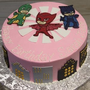 The CB-009G is a round pink birthday cake featuring three PJ Masks characters on top, cityscape designs on the sides, and "Happy Birthday Sarah Jane" written in yellow icing.