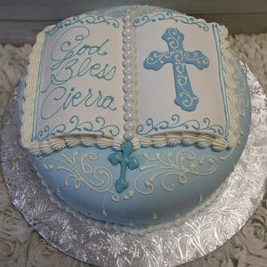 The RC-096 is a round cake designed as an open book with blue and white icing, a cross, and "God Bless Cierra" written on one page. It sits on a silver floral-patterned board.