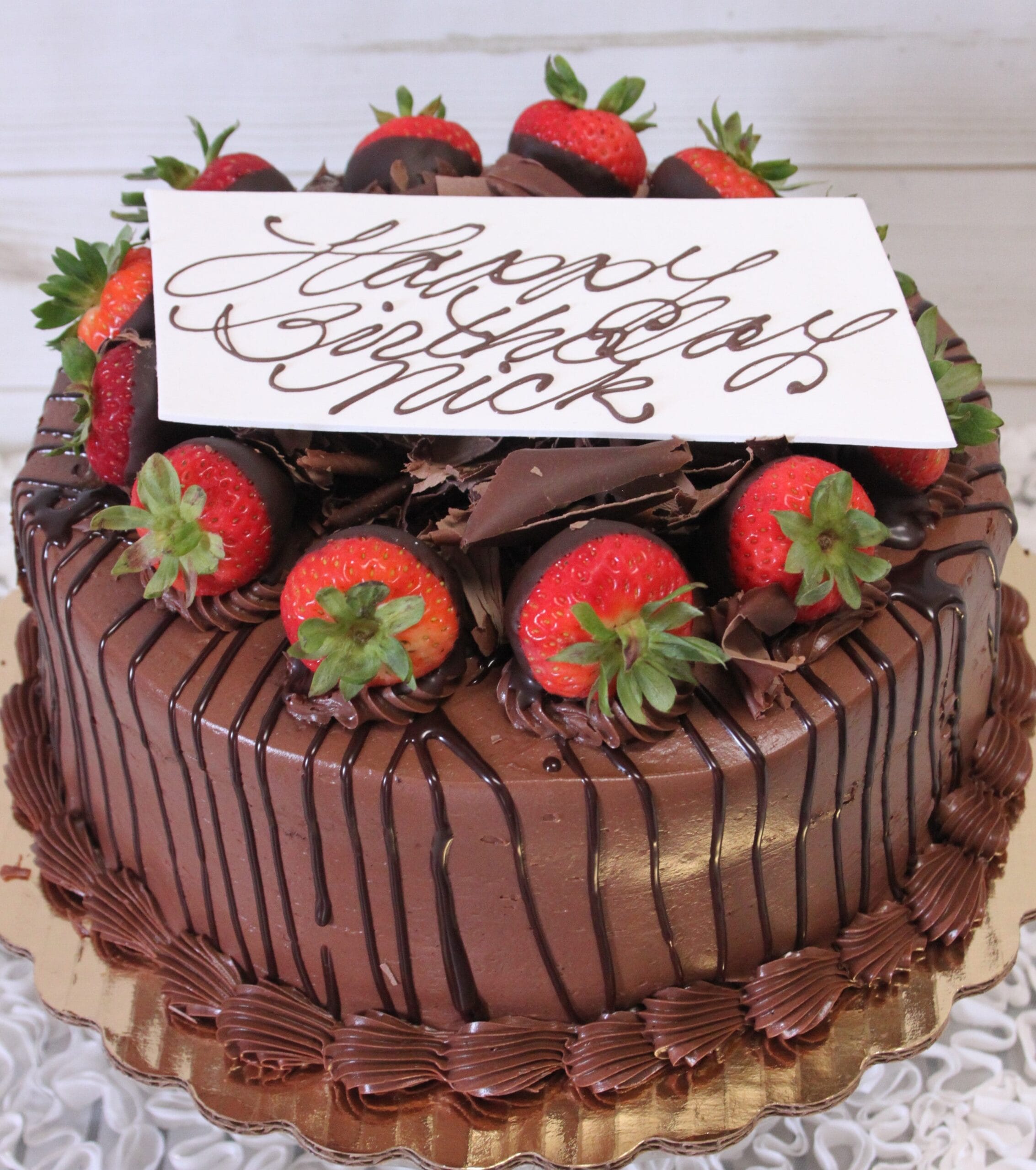 The BD-014A Chocolate/Chocolate is a round chocolate cake garnished with chocolate-dipped strawberries, chocolate shavings, and a white “Happy Birthday” card, presented on a gold cake board.