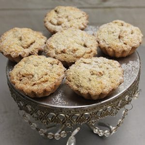 Six Mini Apple Tarts with crumb topping are displayed on an ornate silver cake stand with crystal details, set against a light wooden background.