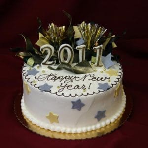 The NY-001 cake is round and decorated with yellow, gray, and purple stars, gold trim, "Happy New Year!" icing, 2011 candles, and gold party decorations on top against a dark red background.