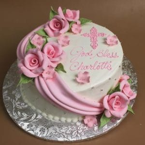 RC-044 is a heart-shaped cake adorned with pink fondant roses, a pink cross, and “God Bless Charlotte” written in pink icing, presented on a silver floral-patterned base.