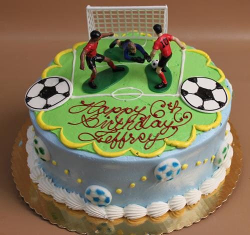 The SC-054 Soccer cake is a round birthday cake topped with a soccer field, two players, a goalie, and a goal. "Happy 6th Birthday Jeffroy" is written on it, with soccer ball designs decorating the sides.