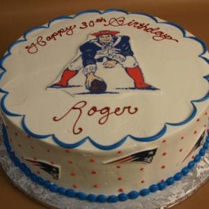 Round cake with white icing, blue border, and New England Patriots logo on top. “Happy 30th Birthday Roger” in red text. Sides feature small Patriots logos and red dots. Product: ST-001 Patriots.