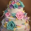 The TC-143 is a three-tiered white cake decorated with colorful fondant flowers and leaves in pink, teal, yellow, and purple, topped with a decorative number 15 on a brown background.