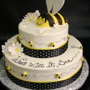 The TC-145 is a two-tier white cake with daisies, small bees, black polka dot ribbon, a fondant bee topper, and "What Will it Bee?" written in cursive on the side.