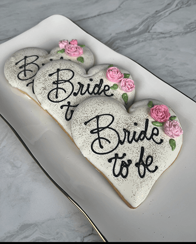 Three heart-shaped sugar cookies from CE-003f bridal, decorated with white icing, pink flowers, and "Bride to be" in black icing, arranged on a rectangular white plate with a gold rim.