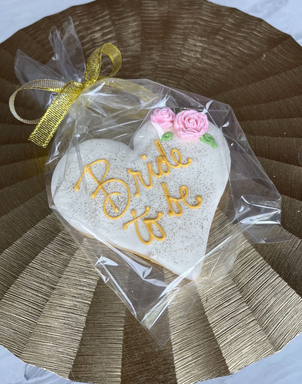 The CE-003g bridal is a heart-shaped cookie with "Bride to be" in yellow icing, pink floral decorations, wrapped in clear plastic with a gold ribbon, and placed on a textured gold placemat.