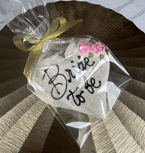 The CE-003h bridal cookie is heart-shaped, decorated with pink roses and "Bride to be" in black icing, wrapped in clear plastic with a gold ribbon, and displayed on a shiny textured gold surface.