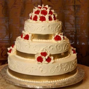The WC-031 is a three-tiered white wedding cake with intricate icing, gold ribbon bands on each layer, and red and white rose toppers, displayed on a silver base.