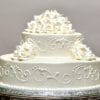WC-032 is a two-tier white cake adorned with detailed piped icing and clusters of white icing roses on the top and sides, presented on a silver tray.