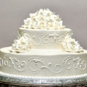 WC-032 is a two-tier white cake adorned with detailed piped icing and clusters of white icing roses on the top and sides, presented on a silver tray.