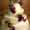 Product WD-076: A three-tiered white wedding cake adorned with purple and white orchids, green leaves, and elegant swirling icing on a silver board, set against a brown background.