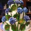 WD-133 is a three-tiered white cake adorned with realistic blue fondant hydrangea flowers and large green leaves, artfully wrapping around each layer.