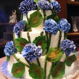 WD-133 is a three-tiered white cake adorned with realistic blue fondant hydrangea flowers and large green leaves, artfully wrapping around each layer.