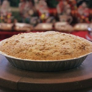 A Pie-Apple with a golden, crumbly topping rests in a foil pan on a tabletop, set against a blurred festive background with red and holiday decorations.