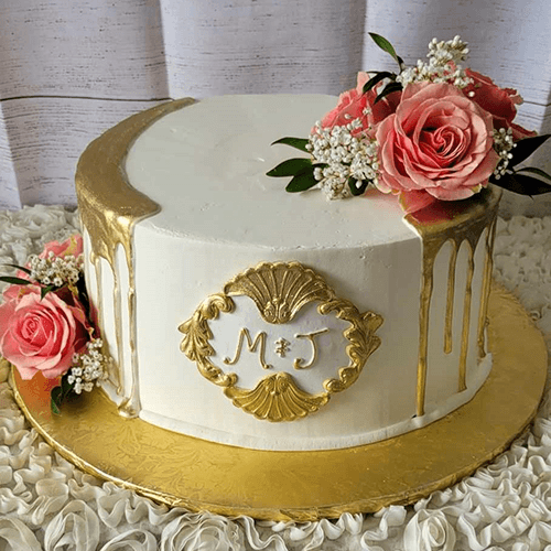 The BR-002 is a round white cake with gold accents, a gold drip design, and an M & J gold plaque. Decorated with pink roses and baby's breath, it sits on a gold cake board.