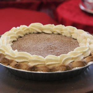 A Pie-Pumpkin with a golden-brown crust is finished with whipped cream swirls and a sprinkle of cinnamon or nutmeg on top, presented on a dark surface with a red cloth in the background.