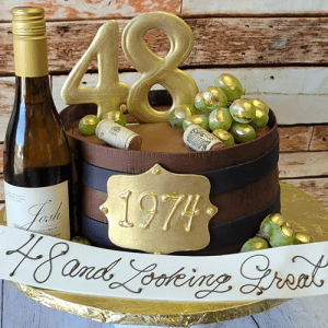 A cake shaped like a wooden wine barrel with gold grapes, wine corks, a “1974” plaque, and big gold “48” toppers sits beside a BD-024T White wine bottle. A banner says, “48 and Looking Great!”.
