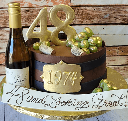 A cake shaped like a wooden wine barrel with gold grapes, wine corks, a “1974” plaque, and big gold “48” toppers sits beside a BD-024T White wine bottle. A banner says, “48 and Looking Great!”.