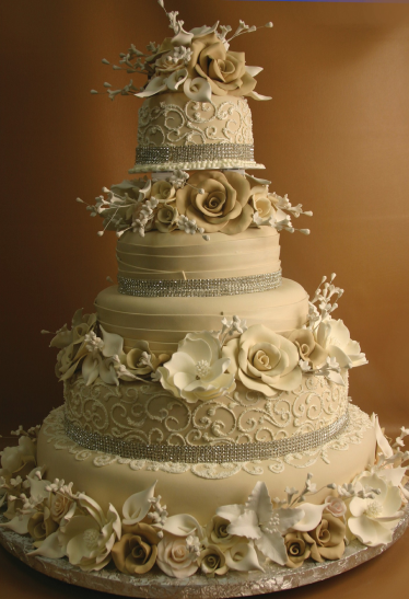 A five-tiered beige wedding cake decorated with intricate white and beige floral designs, large sugar roses, piped patterns, and bands of small silver beads, set against a matching beige background.