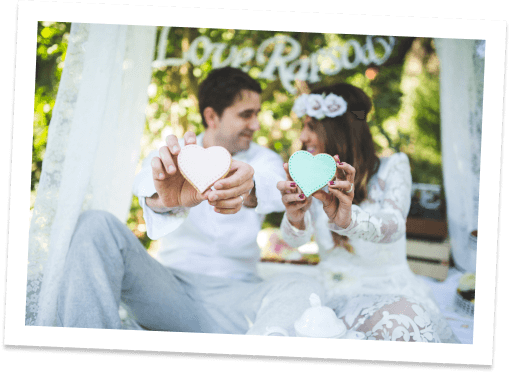 A couple sits outdoors, smiling and holding up pastel heart-shaped cookies. They are dressed in white, with the woman wearing a flower crown. The background is decorated with greenery and a white Love sign.