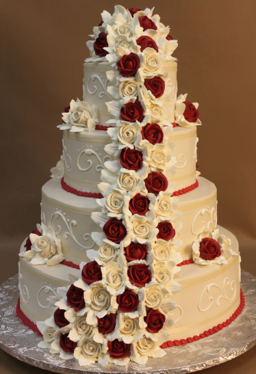 A four-tiered round wedding cake decorated with white frosting, intricate swirl designs, and a cascading arrangement of red and white roses with white lilies. The cake sits on a silver patterned board.