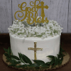 The RC-001I is a white frosted cake adorned with small white flowers, green leaves at the base, a gold cross, and a glittery gold "God Bless" topper, displayed against a wooden background.