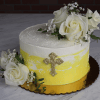 The RC-001J is a round cake with white and yellow frosting, adorned with white roses, baby’s breath, and a gold cross on the side. It sits on a gold cake board over a marble surface.