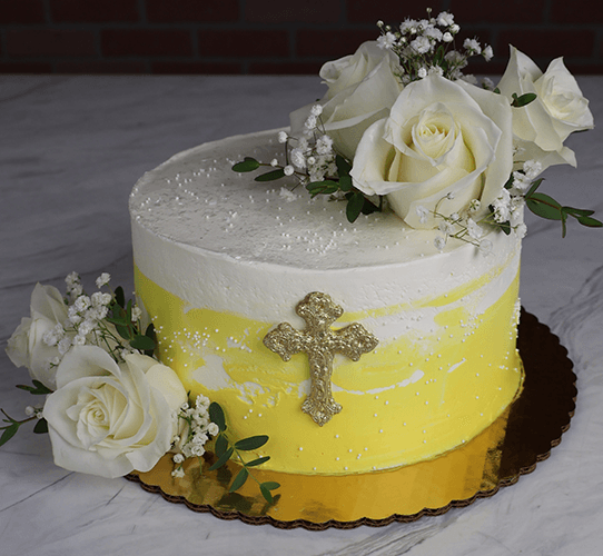 The RC-001J is a round cake with white and yellow frosting, adorned with white roses, baby’s breath, and a gold cross on the side. It sits on a gold cake board over a marble surface.