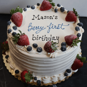 The CB-023I is a white frosted cake topped with strawberries and blueberries, featuring “Masons berry first birthday” in black and blue icing, and presented on a gold board.