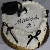 A heart-shaped white cake (GC-0008) with black bows, a black graduation cap, a diploma, and “Mastered it!” in black icing on top. The cake sits on a gold board against a marble background.