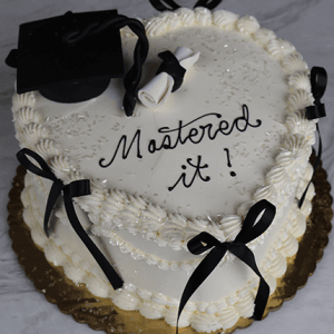 A heart-shaped white cake (GC-0008) with black bows, a black graduation cap, a diploma, and “Mastered it!” in black icing on top. The cake sits on a gold board against a marble background.