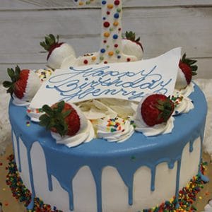 CB-023E: A white cake with blue icing drip, topped with strawberries, colorful sprinkles, and a large 1. Includes a white card inscribed "Happy Birthday Henry" in blue icing.