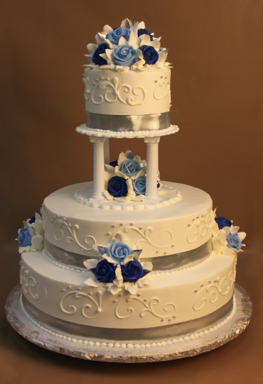 A two-tier white wedding cake with silver ribbons, decorative white icing, and blue and white sugar flowers. The upper tier is supported by white columns and has a floral topper.