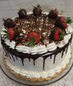 A round Konditor Meister chocolate cake with white frosting, chocolate ganache dripping down the sides, topped with chocolate-covered strawberries, whipped cream, and nuts, displayed on a glass cake stand.