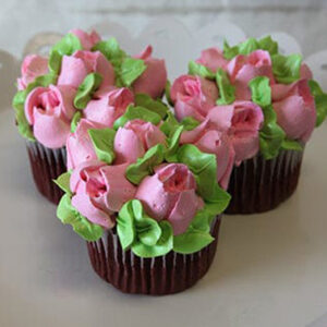 A CC-091 Gold or Chocolate cupcake with white filling is displayed on a white scalloped-edge plate, decorated with colorful buttercream flowers—pink petals—and green leaves.