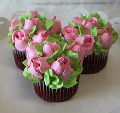 A CC-091 Gold or Chocolate cupcake with white filling is displayed on a white scalloped-edge plate, decorated with colorful buttercream flowers—pink petals—and green leaves.