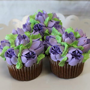 A CC-091 Gold or Chocolate cupcake with white filling is displayed on a white scalloped-edge plate, decorated with colorful buttercream flowers—purple petals—and green leaves.